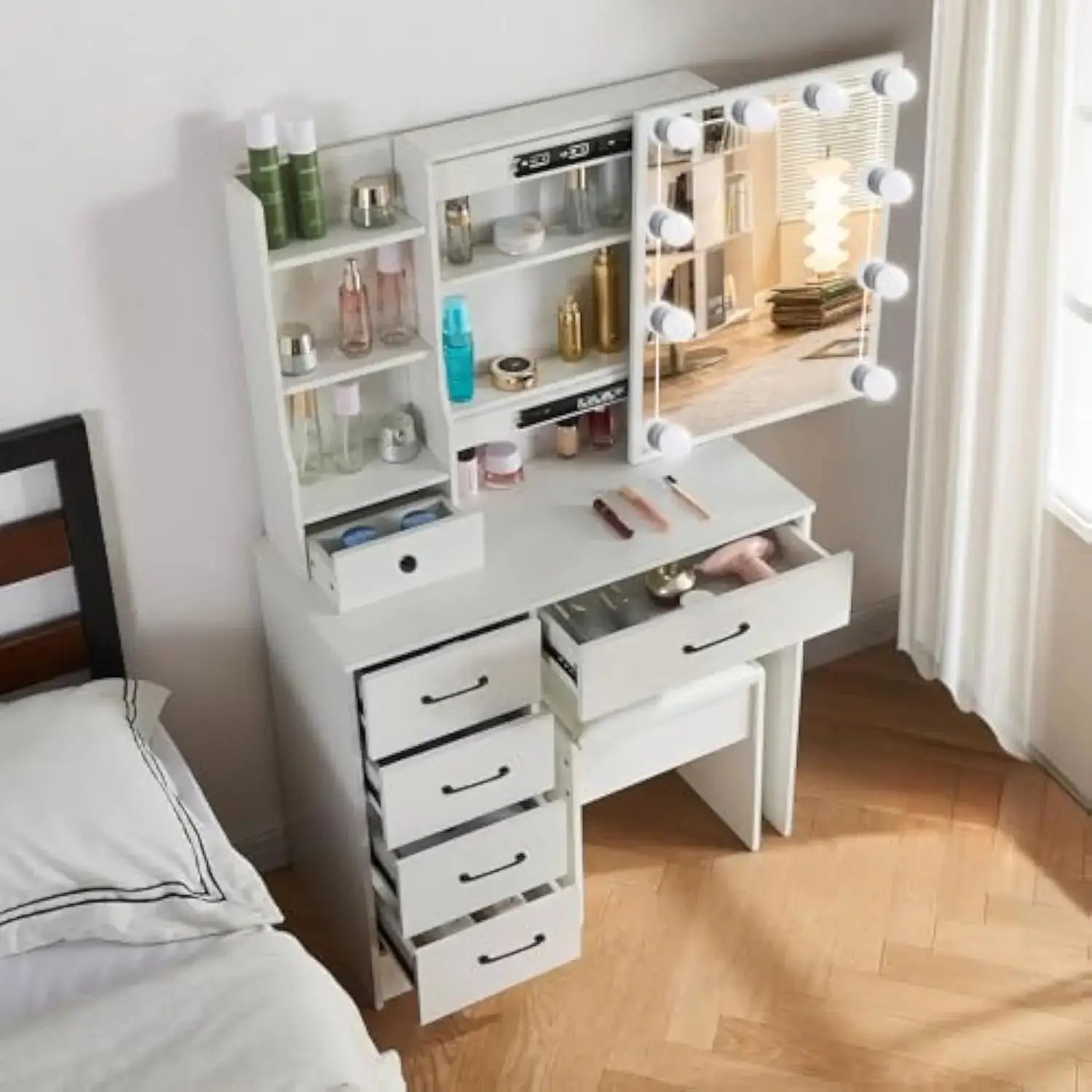 Top view of white makeup vanity desk with lighted mirror, open drawers, and cushioned stool placed beside a bed in a bright bedroom.