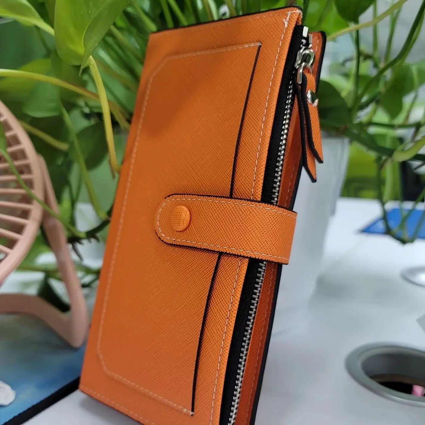 Close-up of orange women's PU leather bifold long wallet with buckle strap and zippered edge, placed near green plants.