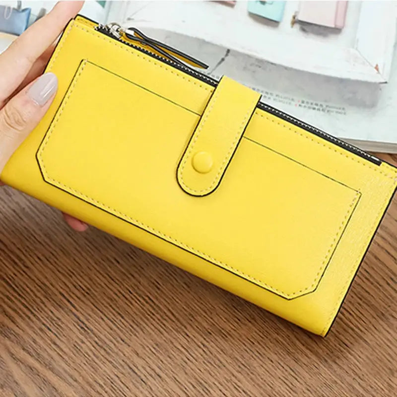 Yellow women's PU leather bifold long wallet with buckle closure and zippered pocket, held in hand on a wooden table.