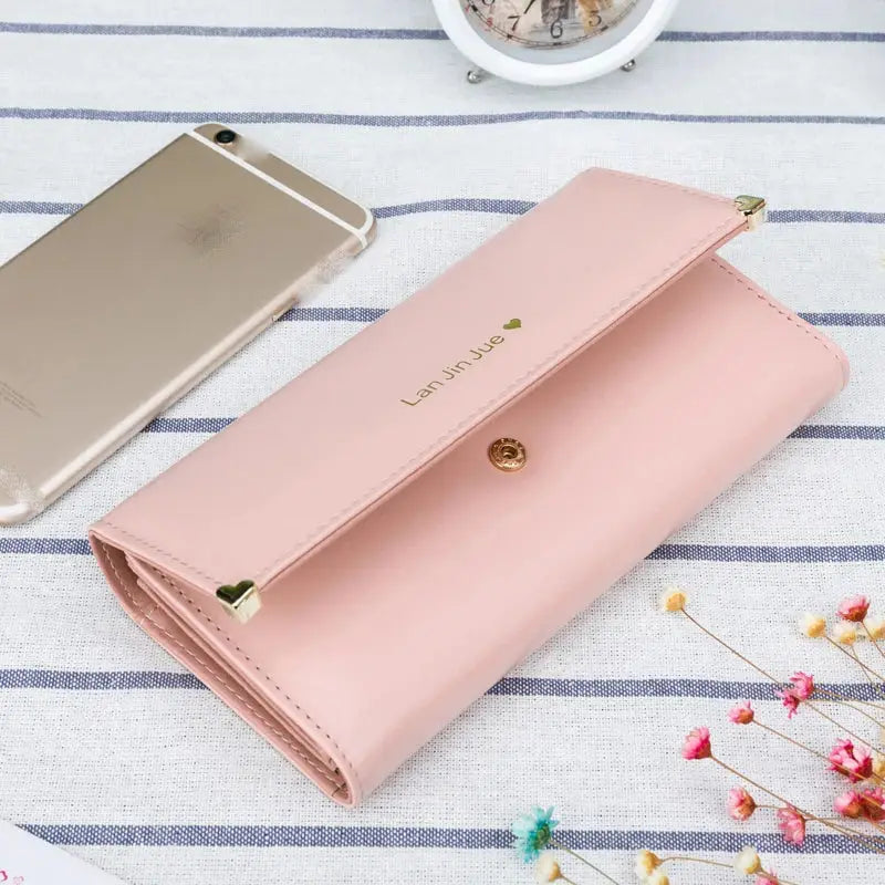 Light pink Women's Long PU Leather Trifold Wallet with snap closure, displayed next to a smartphone on a striped fabric background.
