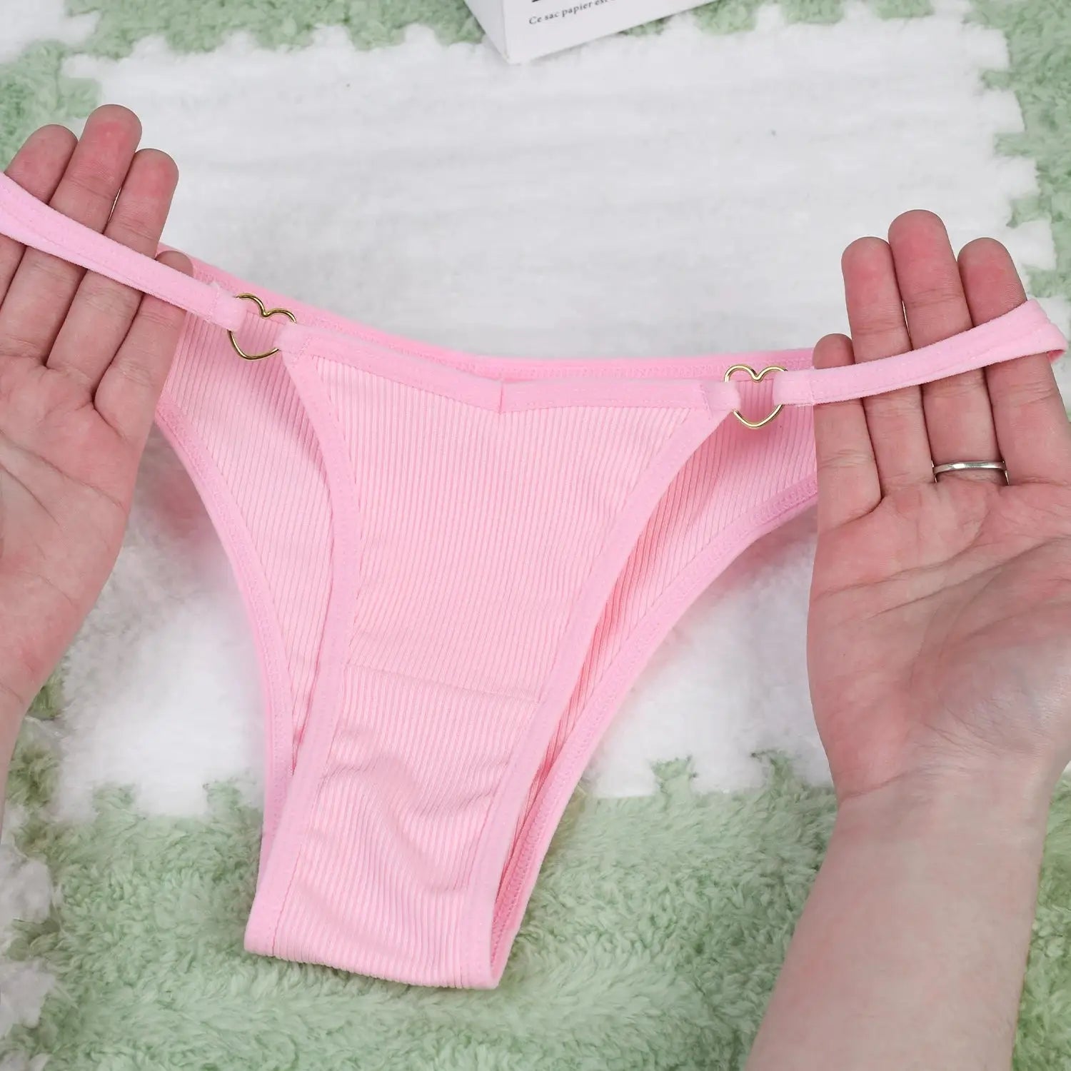Hands holding pink ribbed cotton thong with heart-shaped ring detail, highlighting soft, breathable fabric and low-rise fit.